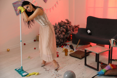 Woman with mop suffering from hangover in messy room after New Year party
