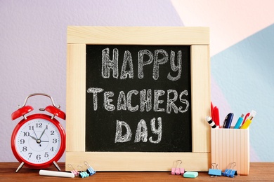 Photo of Little blackboard with inscription HAPPY TEACHER'S DAY, alarm clock and stationery on table