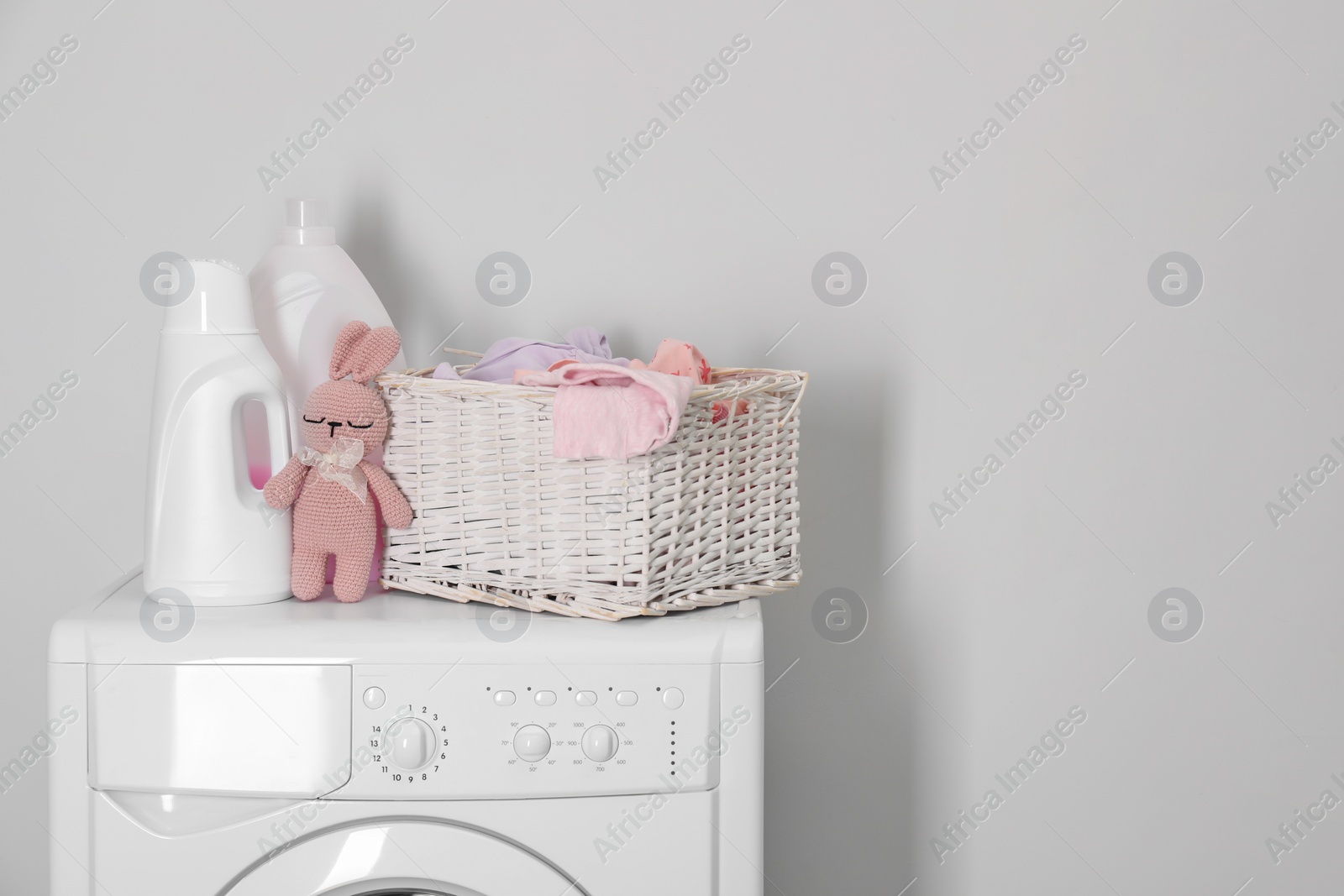 Photo of Baby clothes in wicker basket, laundry detergents and toy bunny on washing machine near light wall, space for text