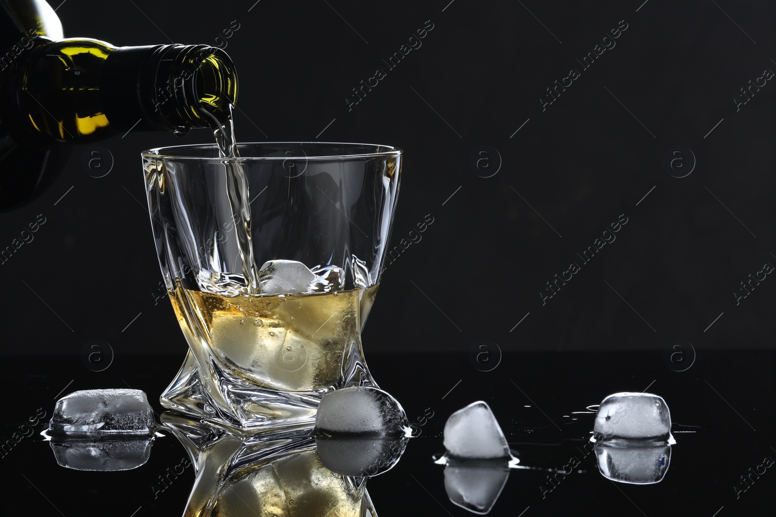 Photo of Pouring tasty whiskey from bottle into glass with ice at mirror table against black background, closeup. Space for text