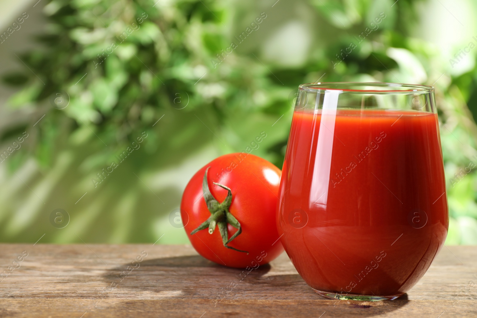 Photo of Delicious juice in glass and fresh tomato on wooden table against blurred background, space for text