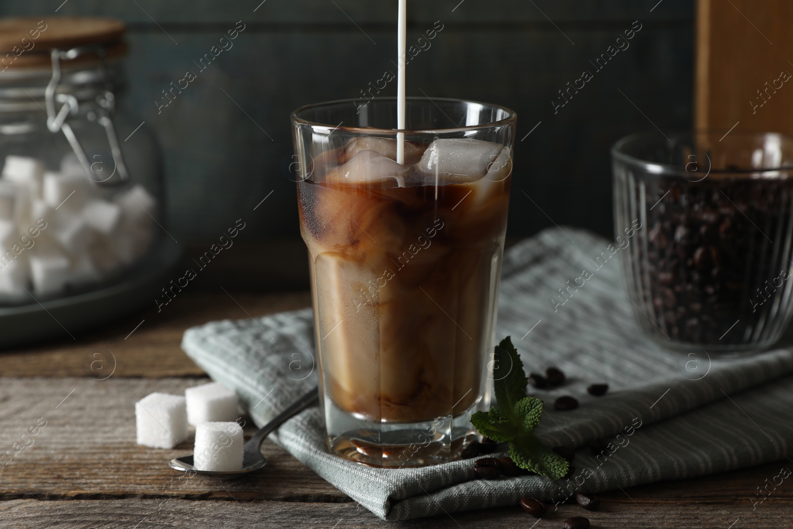 Photo of Pouring milk into glass of delicious iced coffee, mint and sugar cubes on wooden table