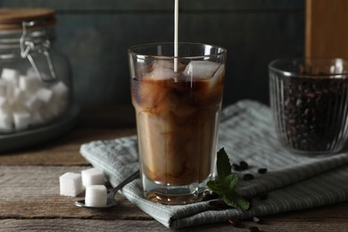 Photo of Pouring milk into glass of delicious iced coffee, mint and sugar cubes on wooden table