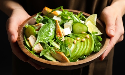 Photo of Woman holding bowl of delicious salad with chicken, arugula and avocado on dark background, closeup