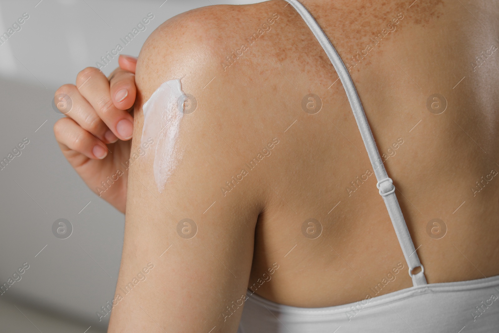 Photo of Woman applying body cream onto shoulder on blurred background, closeup