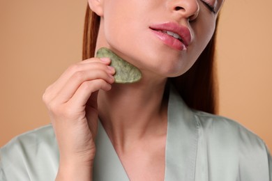 Young woman massaging her face with jade gua sha tool on pale orange background, closeup