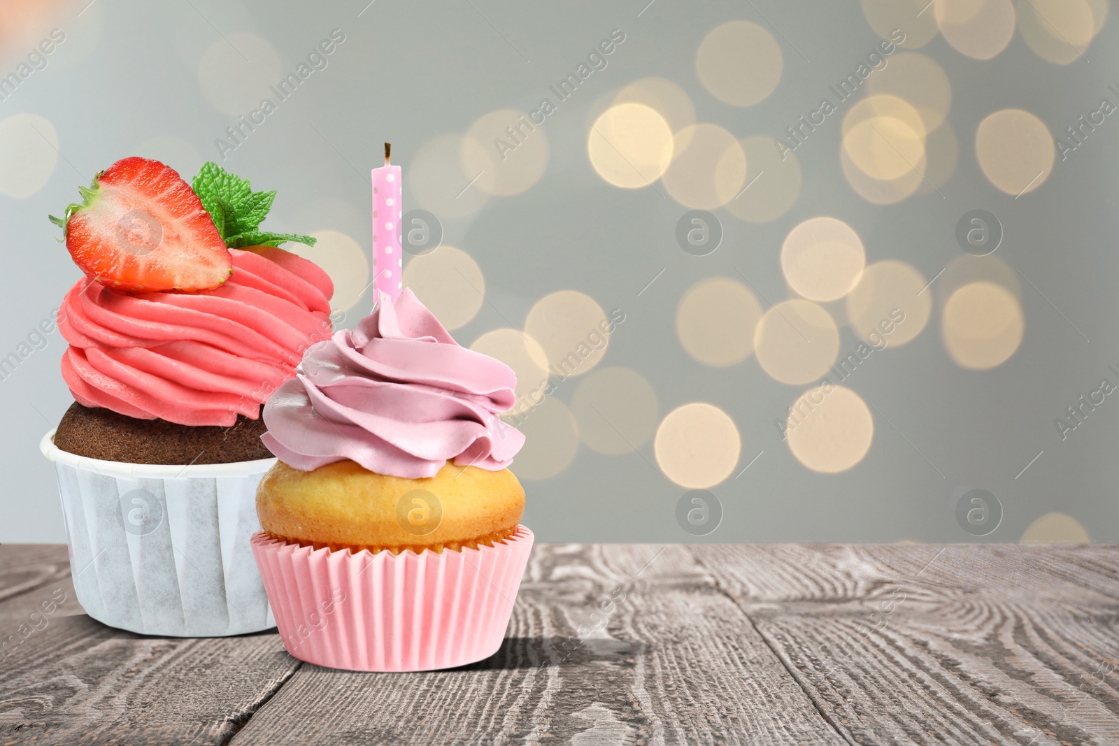 Image of Sweet birthday cupcakes on wooden table against blurred lights. Space for text