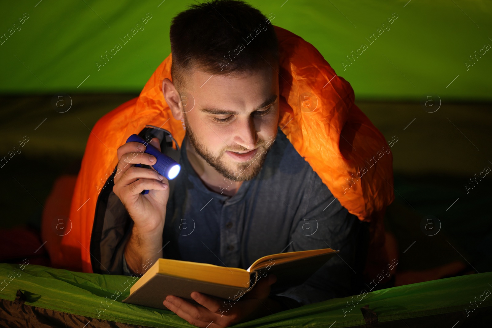 Photo of Young man with flashlight reading book in tent