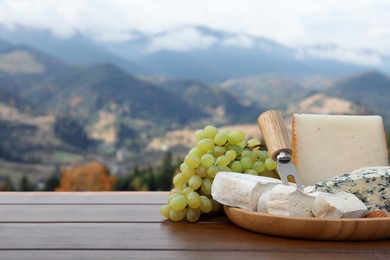 Different types of delicious cheeses, nuts and grapes on wooden table against mountain landscape. Space for text