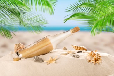 Image of Corked glass bottle with rolled paper note and seashells on sandy beach with palms near ocean