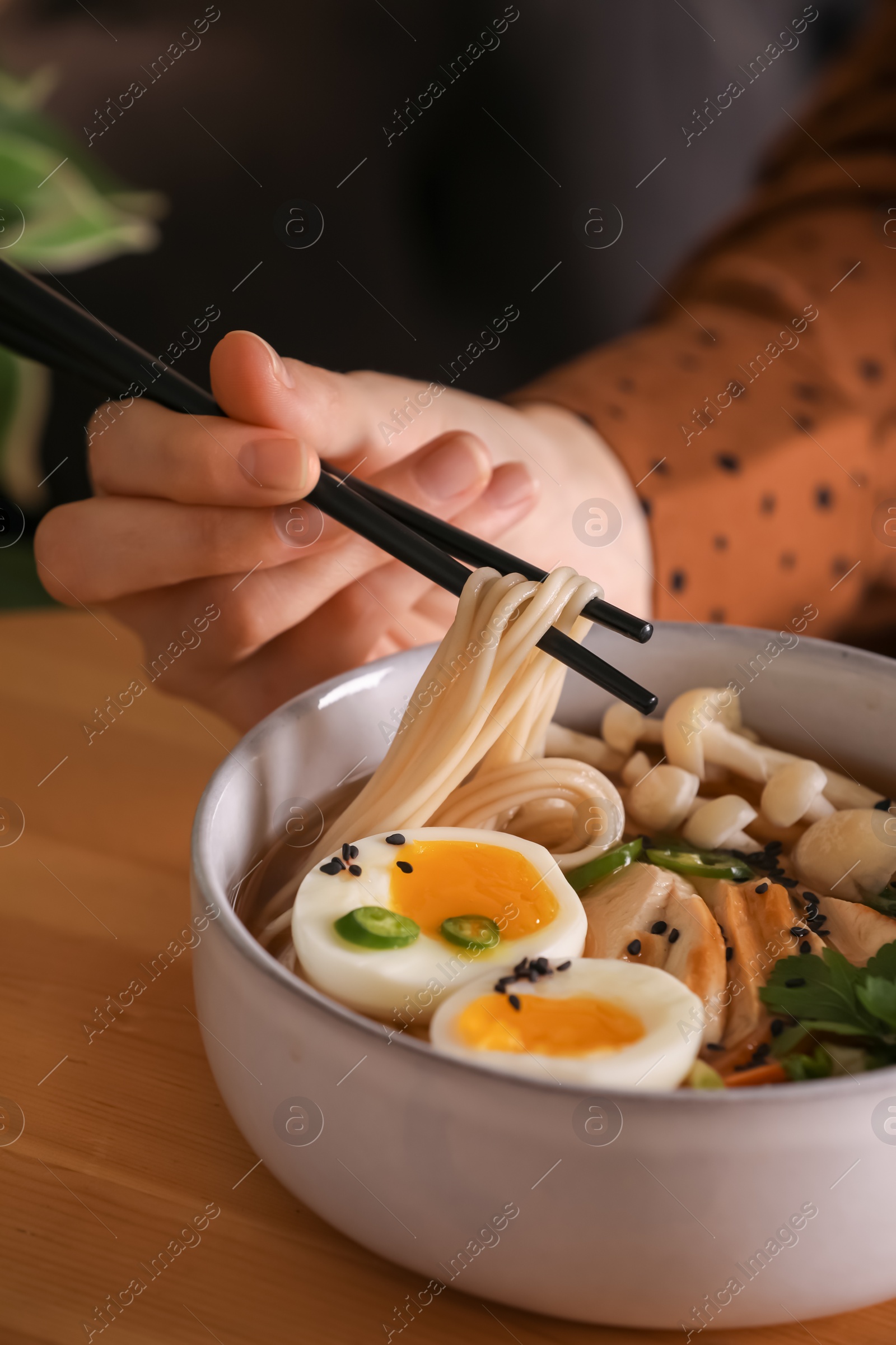 Photo of Woman eating delicious ramen with chopsticks at wooden table, closeup. Noodle soup