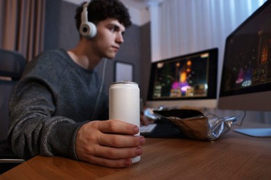 Young man with energy drink playing video game at wooden desk indoors, focus on can