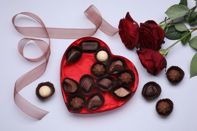 Heart shaped box with delicious chocolate candies, roses and ribbon on white background, flat lay