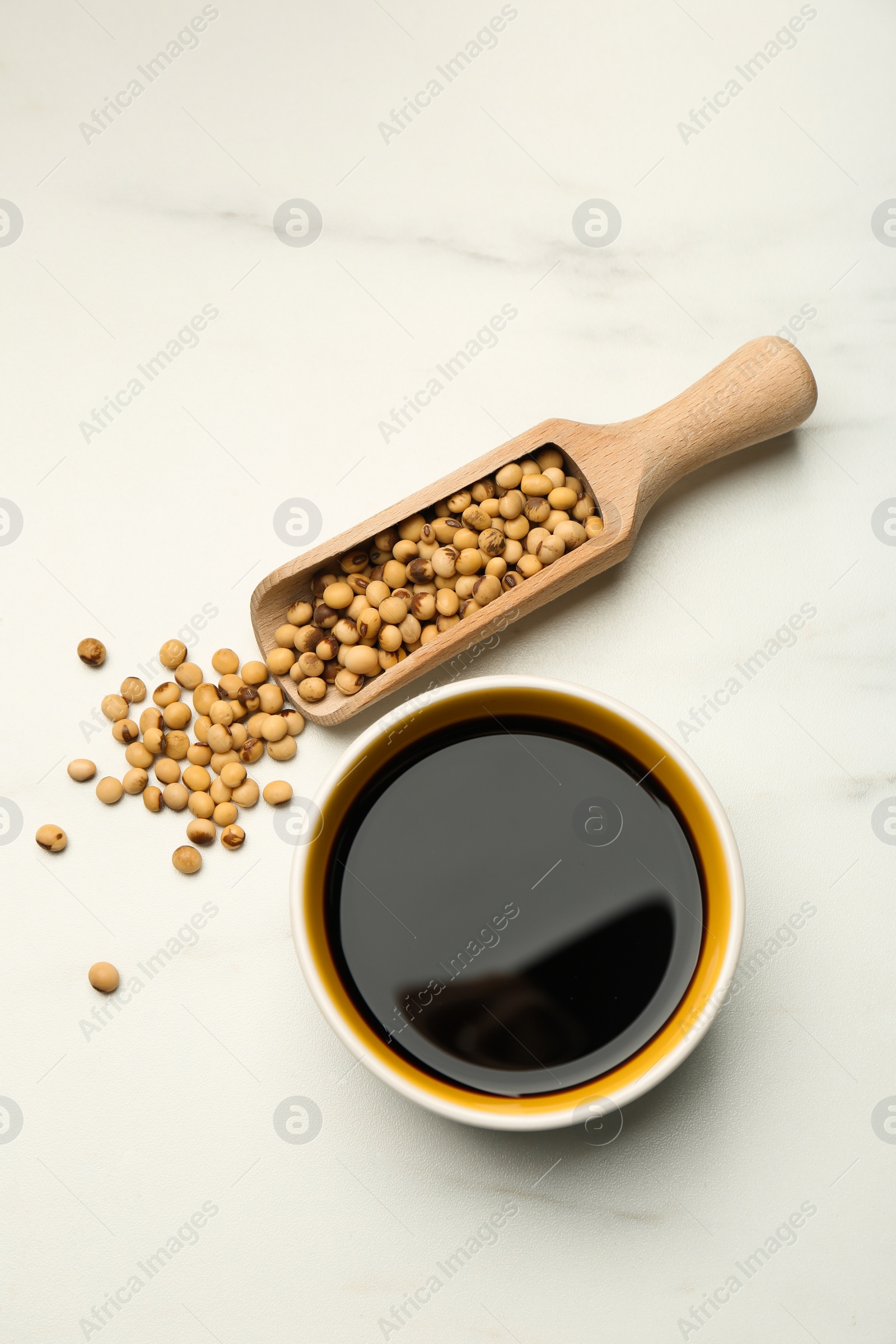 Photo of Soy sauce in bowl and scoop with soybeans on white marble table, flat lay