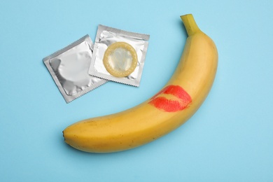 Photo of Condoms and banana with lipstick kiss mark on light blue background, flat lay. Safe sex