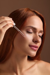 Beautiful young woman applying cosmetic serum onto her face on brown background, closeup