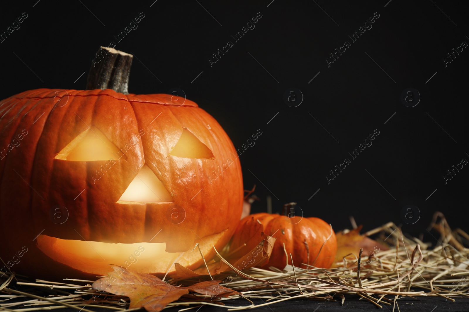 Photo of Pumpkin jack o'lantern, straw and autumn leaves on table in darkness, space for text. Halloween decor