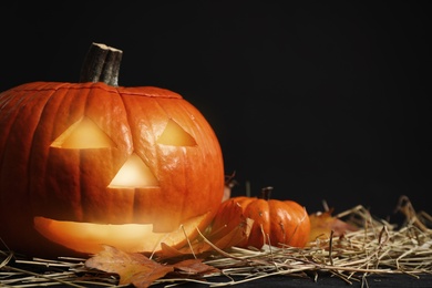 Photo of Pumpkin jack o'lantern, straw and autumn leaves on table in darkness, space for text. Halloween decor