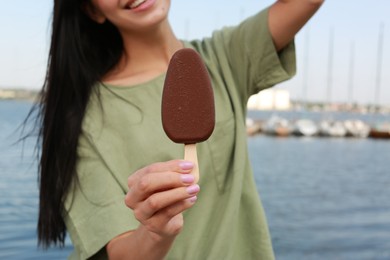 Photo of Young woman holding ice cream glazed in chocolate near river, closeup