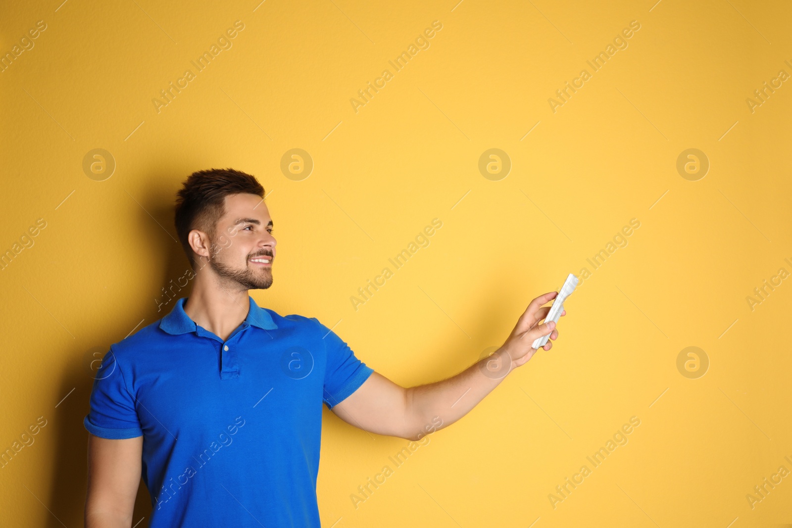 Photo of Happy young man operating air conditioner with remote control on yellow background. Space for text