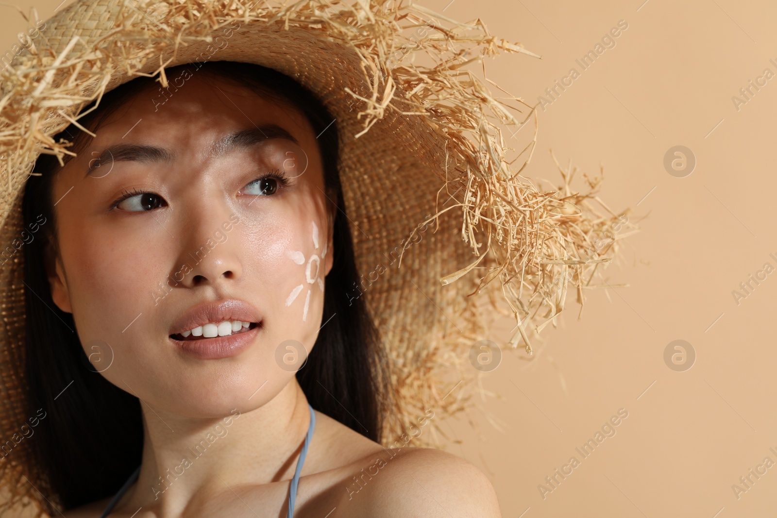 Photo of Beautiful young woman in straw hat with sun protection cream on her face against beige background, space for text