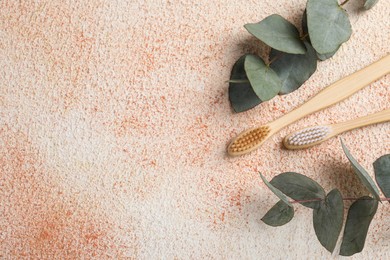 Photo of Bamboo toothbrushes and eucalyptus branches on beige textured table, flat lay. Space for text