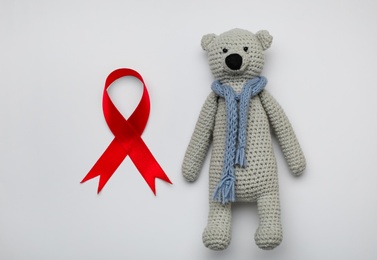Photo of Cute knitted toy bear and red ribbon on light grey background, flat lay. AIDS disease awareness