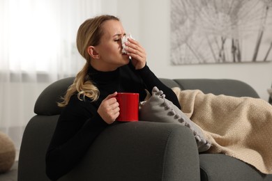 Photo of Sick woman with cup of drink blowing nose in tissue on sofa at home. Cold symptoms