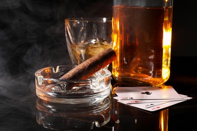 Photo of Smoldering cigar, ashtray, playing cards and whiskey on black mirror surface