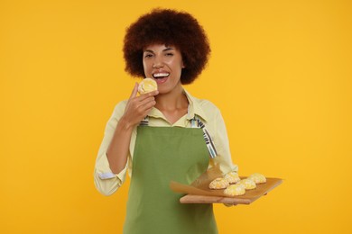 Photo of Happy young woman in apron holding board with cookies on orange background