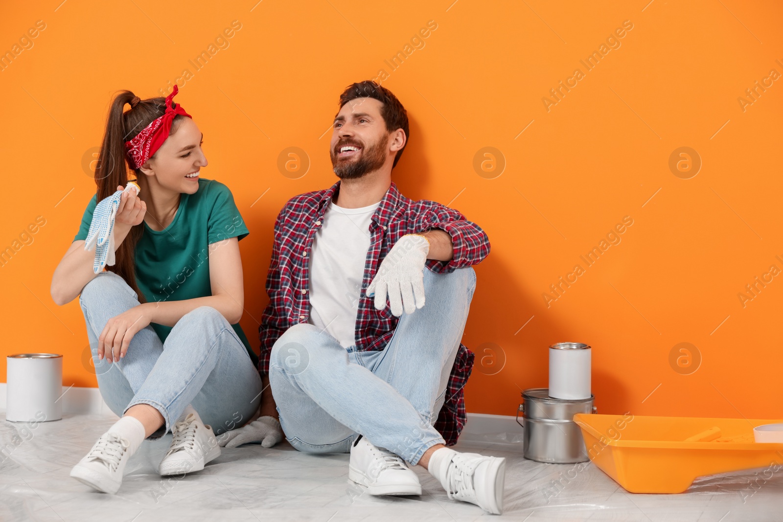 Photo of Happy designers sitting on floor with painting equipment near freshly painted orange wall indoors