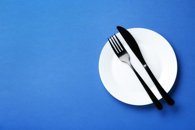 Clean white plate with cutlery on blue background, top view. Space for text