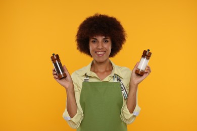 Happy young woman in apron holding shakers with pepper and salt on orange background