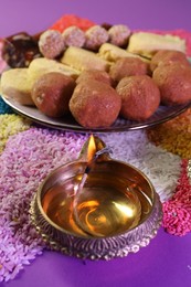 Diwali celebration. Tasty Indian sweets, diya lamp and colorful rangoli on violet table, closeup