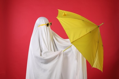 Person in ghost costume and sunglasses holding yellow umbrella on red background