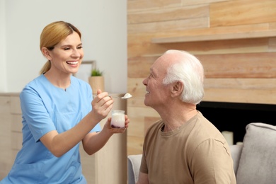 Photo of Nurse feeding elderly man with yogurt indoors. Assisting senior people