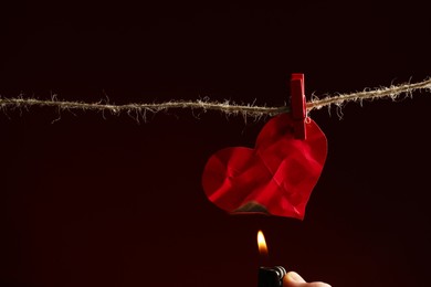 Lighting red paper heart with lighter on rope against burgundy background, space for text. Broken heart