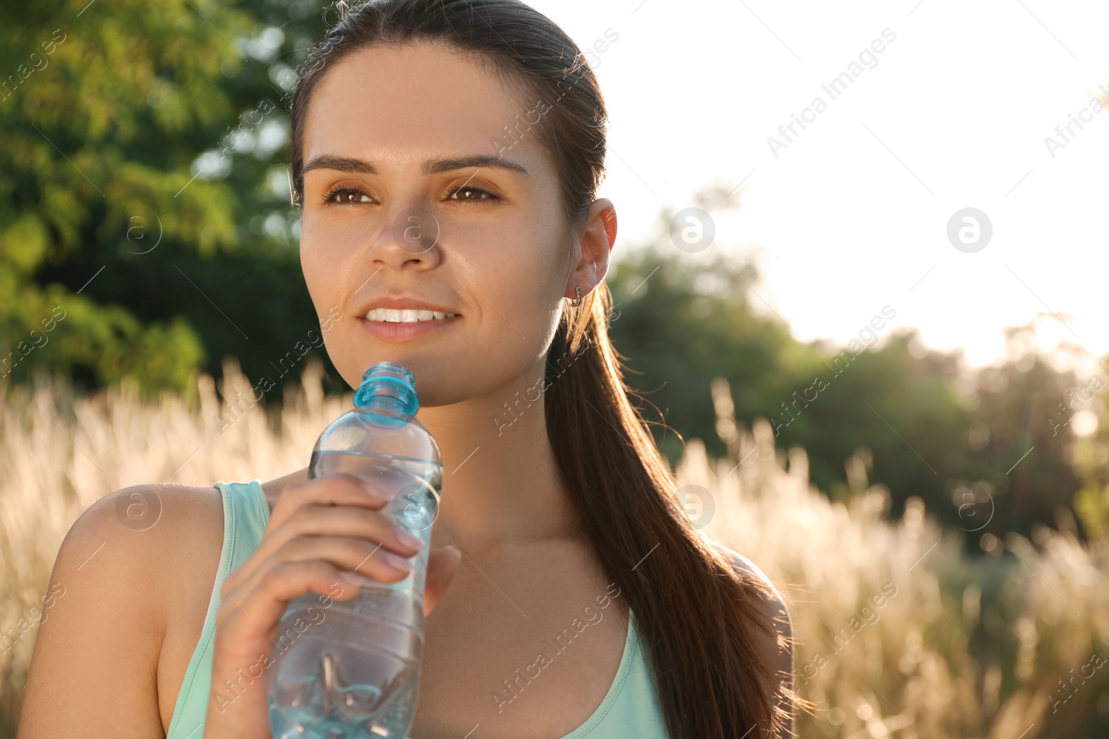 Photo of Happy young woman drinking water outdoors on hot summer day. Refreshing drink