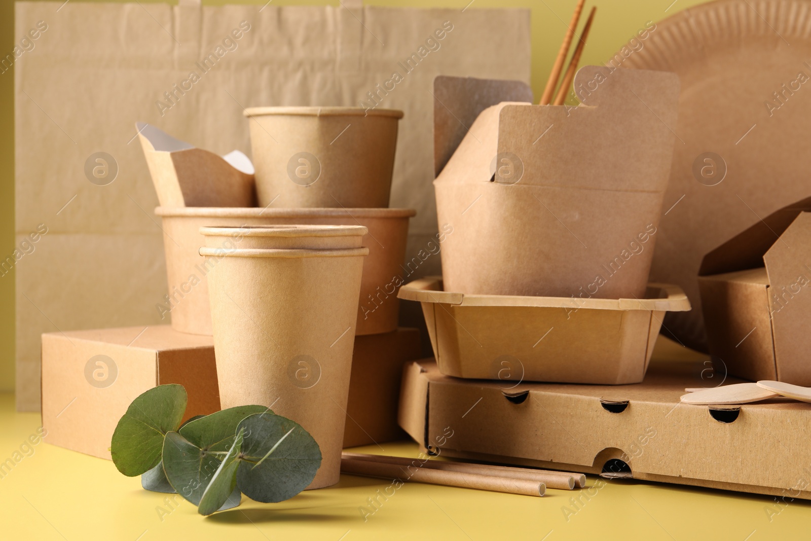 Photo of Eco friendly food packaging. Paper containers, tableware, bag and eucalyptus leaves on pale yellow background