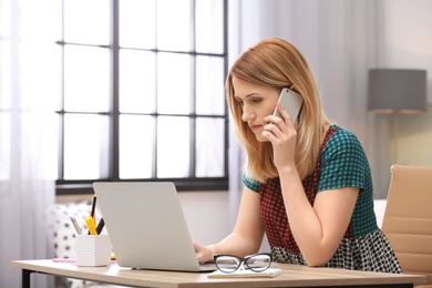 Photo of Young woman talking on phone while working with laptop at desk in home office