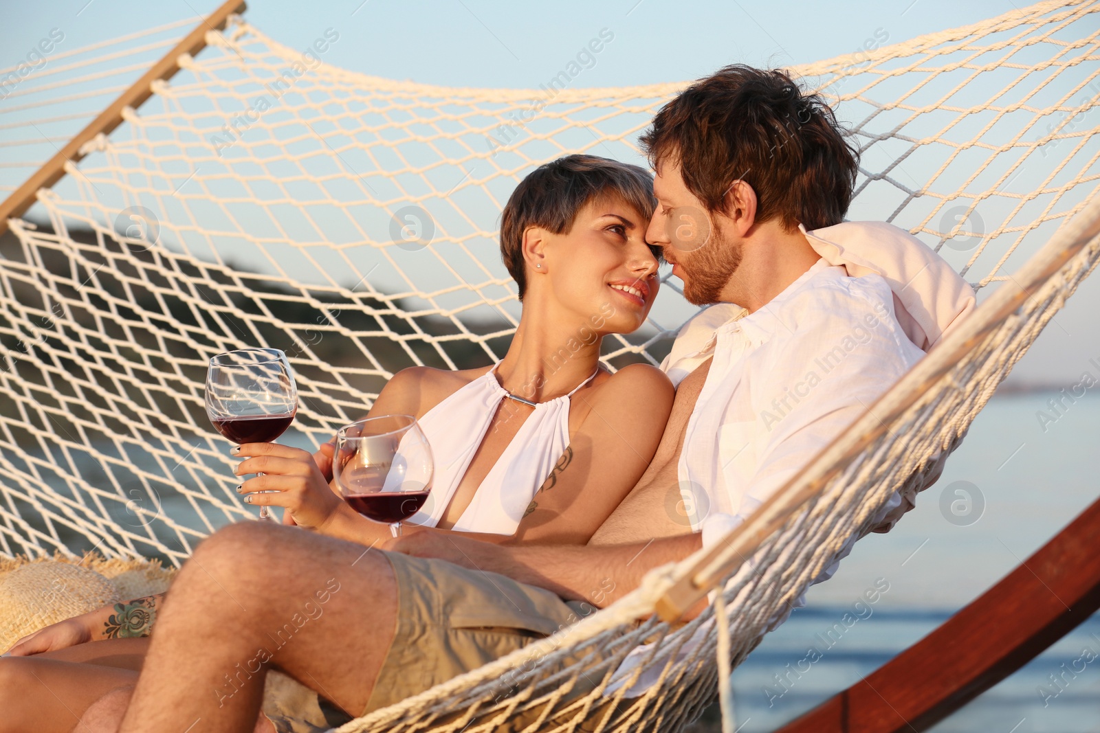 Photo of Young couple resting with glasses of wine in hammock on beach