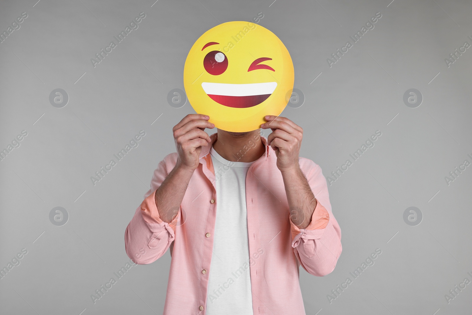 Photo of Man covering face with happy winking emoticon on grey background