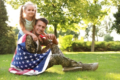 Father in military uniform with American flag and his little daughter sitting on grass at park