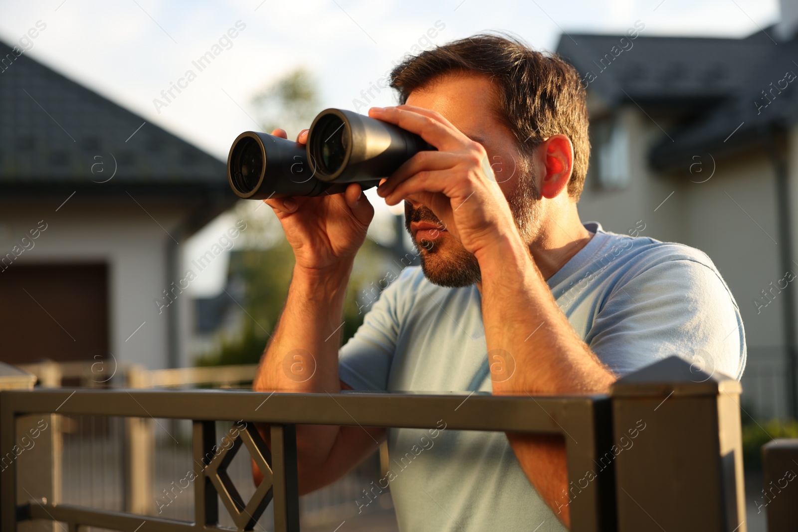 Photo of Concept of private life. Curious man with binoculars spying on neighbours over fence outdoors