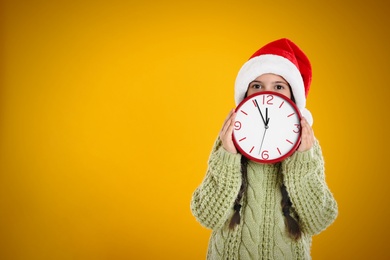 Photo of Girl in Santa hat with clock on yellow background, space for text. Christmas countdown