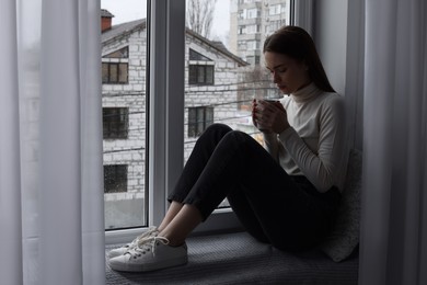 Photo of Melancholic young woman with cup of drink near window indoors, space for text. Loneliness concept