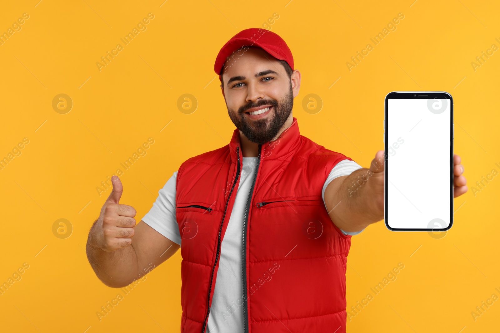 Image of Happy courier holding smartphone with empty screen and showing thumbs up on orange background