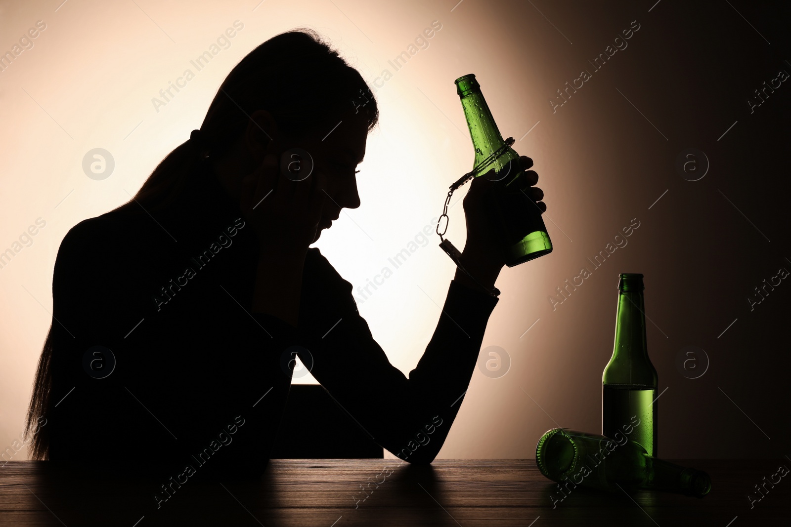 Photo of Alcohol addiction. Silhouette of woman handcuffed with beer bottle at wooden table, backlit