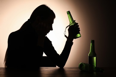 Photo of Alcohol addiction. Silhouette of woman handcuffed with beer bottle at wooden table, backlit