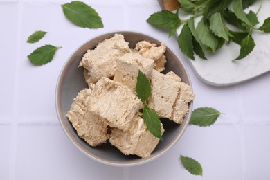 Photo of Bowl with pieces of tasty halva and mint leaves on white tiled table, flat lay